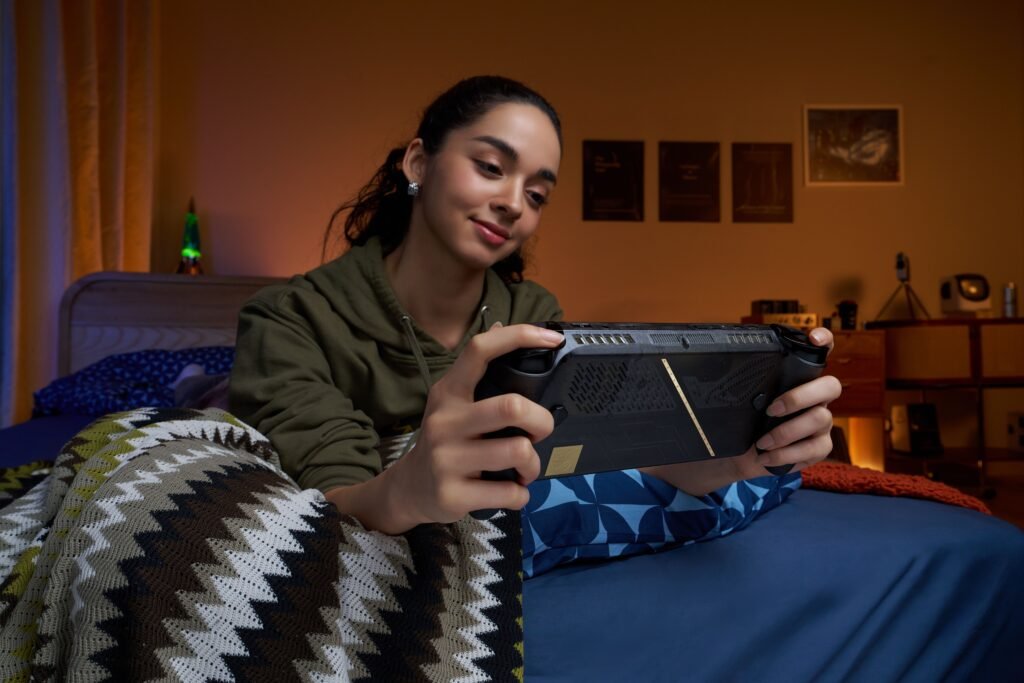 Mujer joven recostada en una cama sosteniendo y jugando con la consola ROG Xbox Ally X20 en acabado translúcido con detalles dorados.